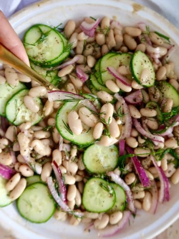 Spoon scooping a portion of the bean salad from the bowl, showcasing white beans, cucumber slices, red onion, and dill.