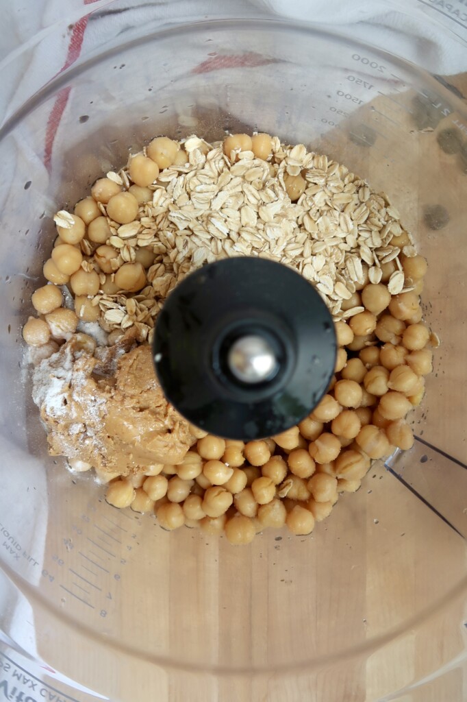 Food processor containing chickpeas, oats, peanut butter, and other ingredients before blending.