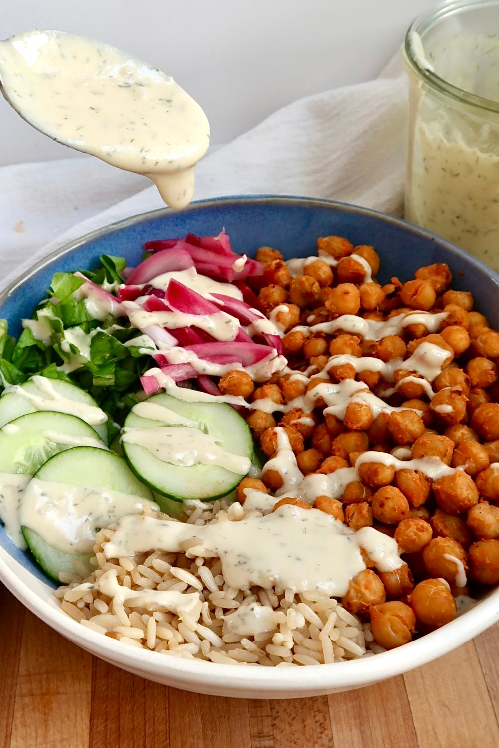 Spoon drizzling dill sauce over buffalo chickpeas, cucumber and brown rice. 