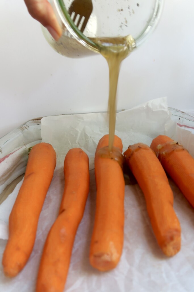 Peeled carrots on a sheet pan with parchment paper with a glaze being poured over top.