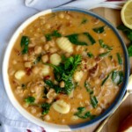 Bowl with creamy gnocchi soup with chickpeas, spinach, sundried tomato on a cutting board with a spoon to the side.