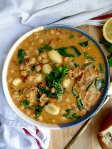 Bowl with creamy gnocchi soup with chickpeas, spinach, sundried tomato on a cutting board with a spoon to the side.