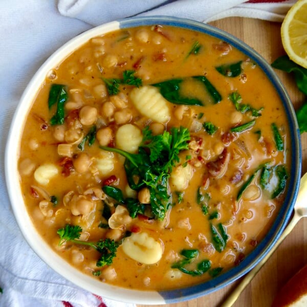 Bowl with creamy gnocchi soup with chickpeas, spinach, sundried tomato on a cutting board with a spoon to the side.