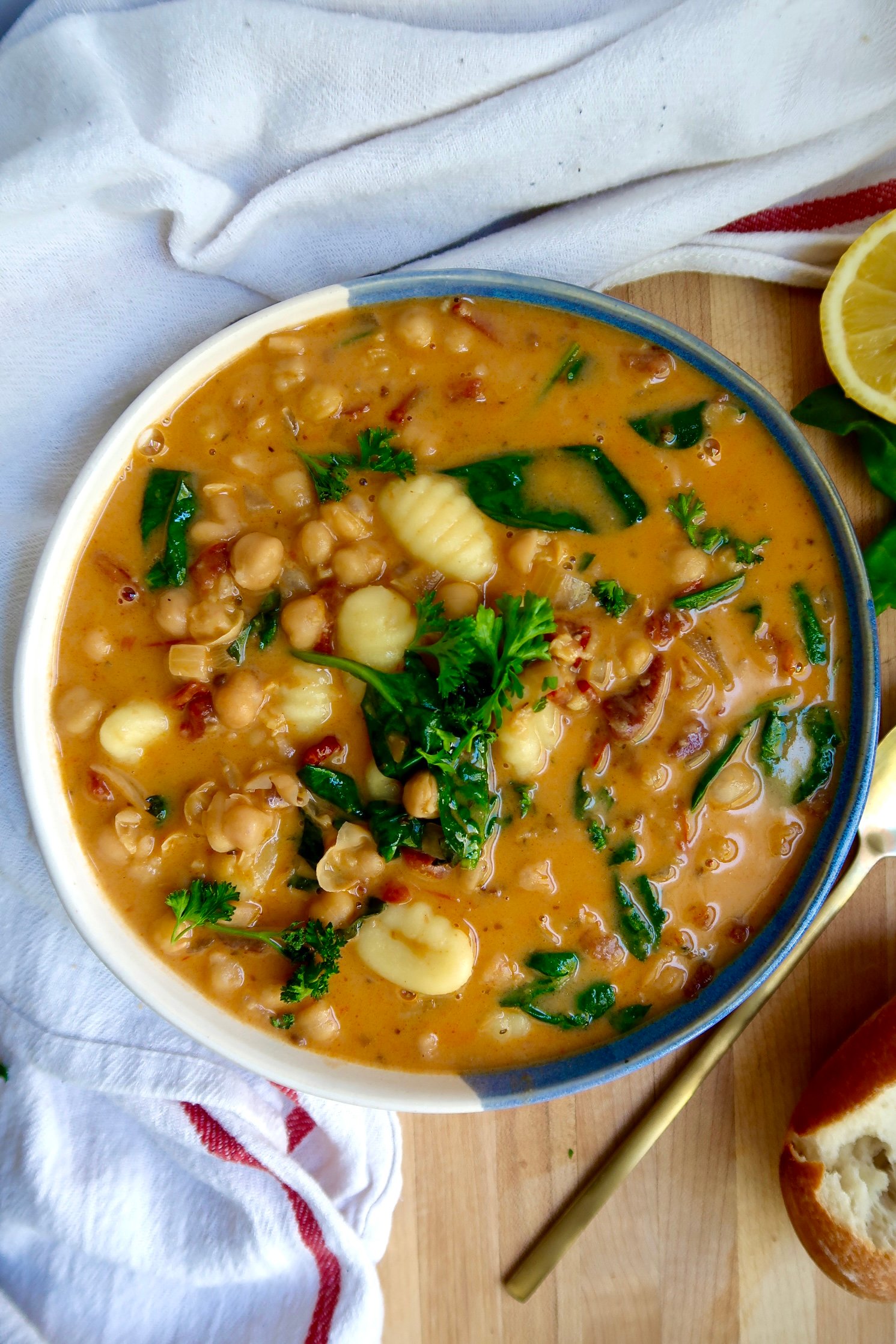 Bowl with creamy gnocchi soup with chickpeas, spinach, sundried tomato on a cutting board with a spoon to the side. 