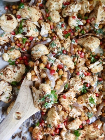 Wooden spoon scooping up some roasted cauliflower and chickpeas as well as pomegranate seeds and red onion with a sauce drizzled on top.
