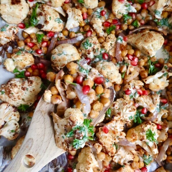 Wooden spoon scooping up some roasted cauliflower and chickpeas as well as pomegranate seeds and red onion with a sauce drizzled on top.