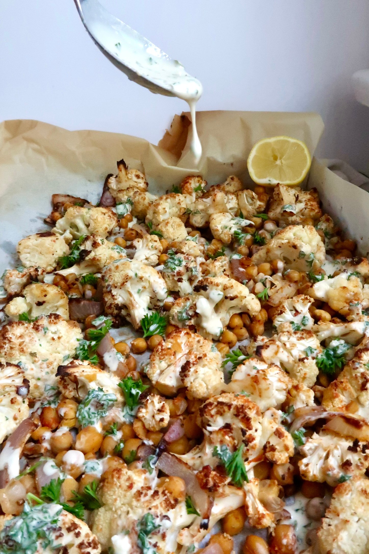 Roasted cauliflower, chickpeas and red onion on a sheet pan with red onion and a creamy sauce being drizzled on top with a spoon.
