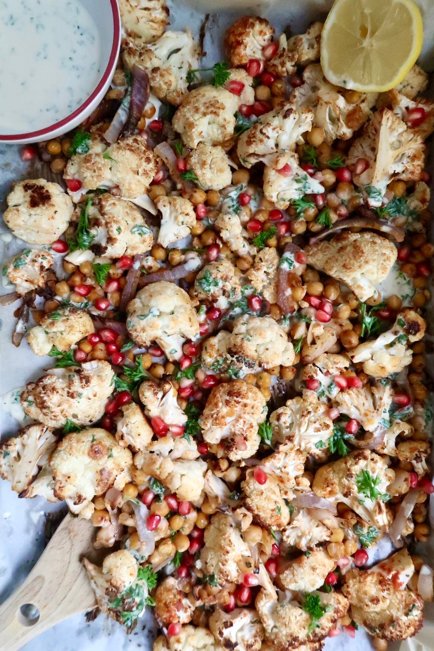 Roasted cauliflower and chickpeas on a sheet pan with a sauce to the side and a sliced lemon, with parsley on top and a creamy sauce drizzled on top.