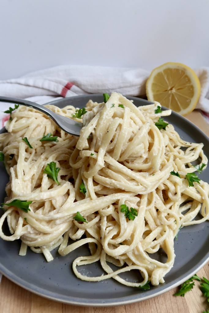 Plate with creamy pasta linquine with parlsey on top and a lemon to the side with a fork spiraling some linguine.