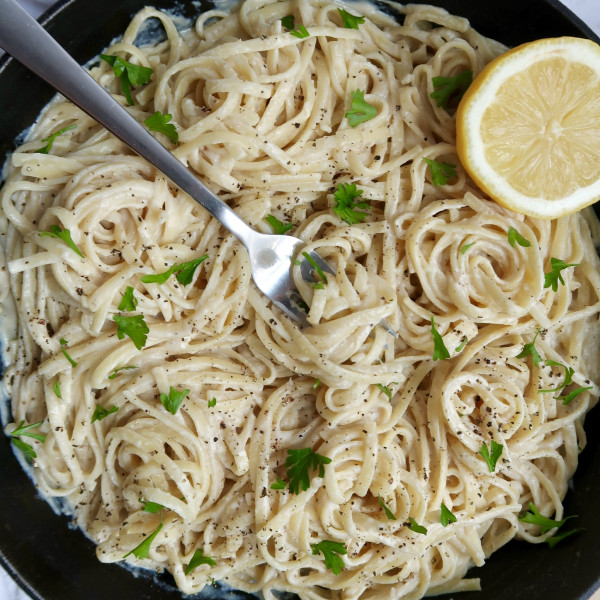 Top down view of cast iron pan with creamy pasta linquine inside with parley and lemon as garnish and a fork spiraling some pasta.