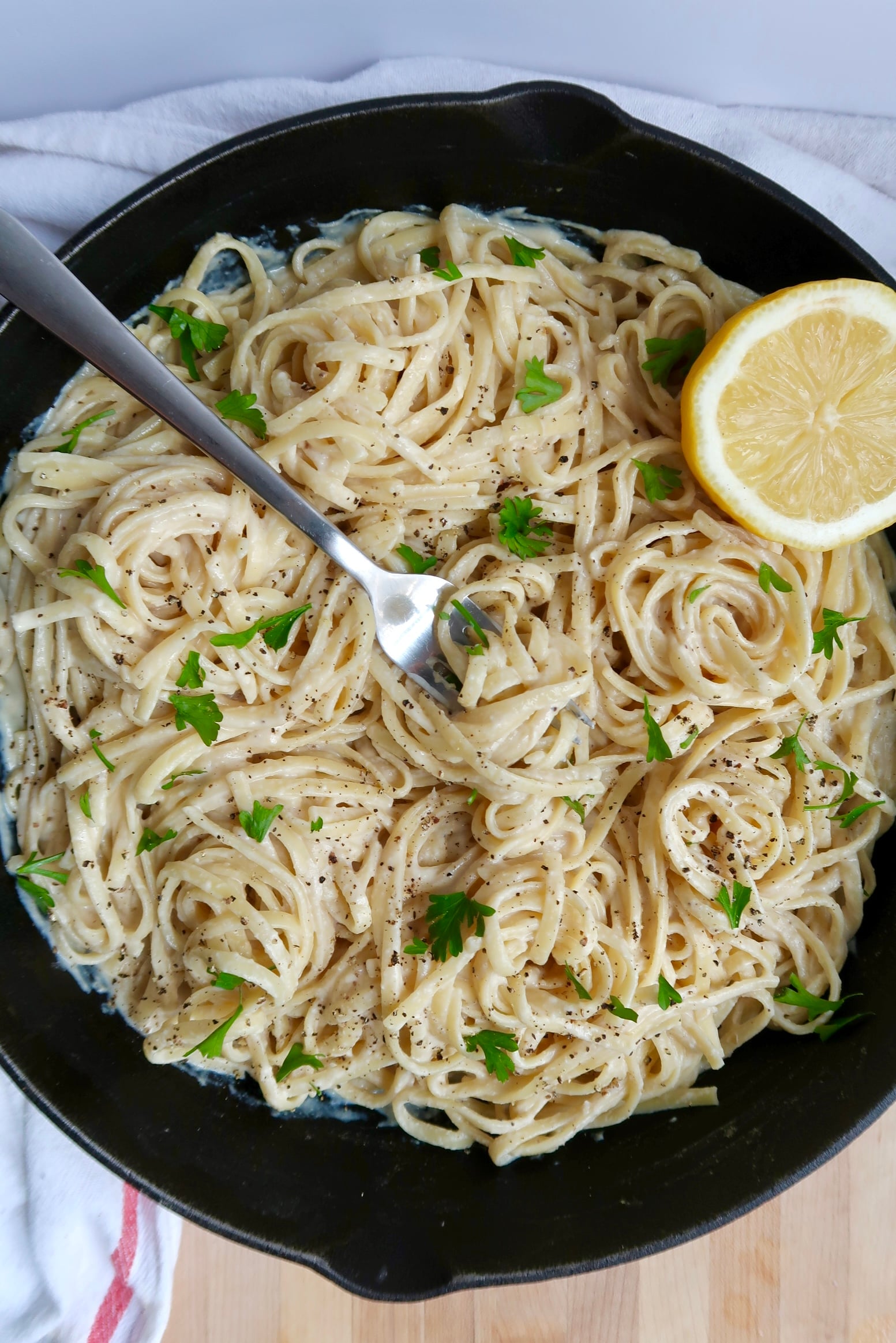 Top down view of cast iron pan with creamy pasta linquine inside with parley and lemon as garnish and a fork spiraling some pasta.