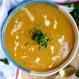 Bowl with creamy pumpkin soup and coconut milk drizzled on top with parlsey and pumpkin seeds on top and a spoon off to the side.