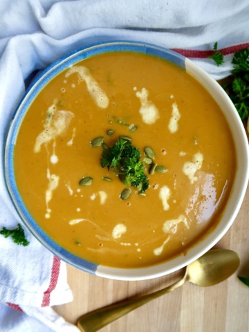 Bowl with creamy pumpkin soup and coconut milk drizzled on top with parlsey and pumpkin seeds on top and a spoon off to the side.