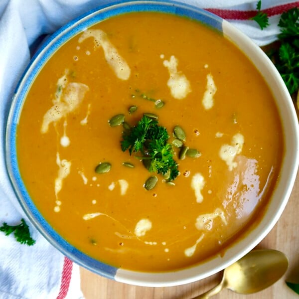 Bowl with creamy pumpkin soup and coconut milk drizzled on top with parlsey and pumpkin seeds on top and a spoon off to the side.