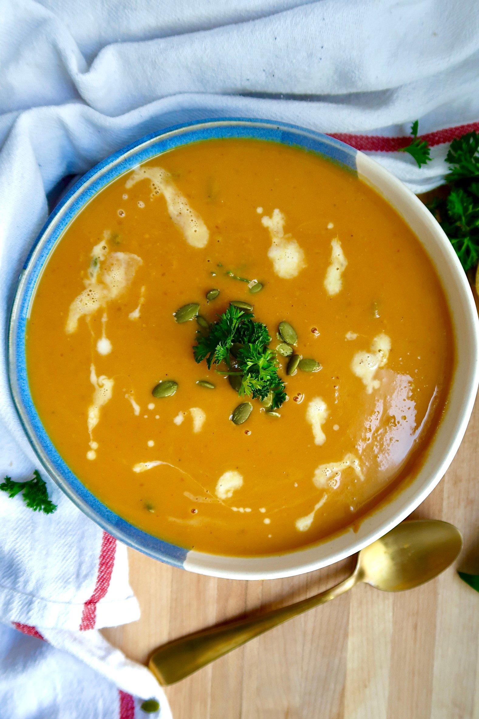 Bowl with creamy pumpkin soup and coconut milk drizzled on top with parlsey and pumpkin seeds on top and a spoon off to the side.