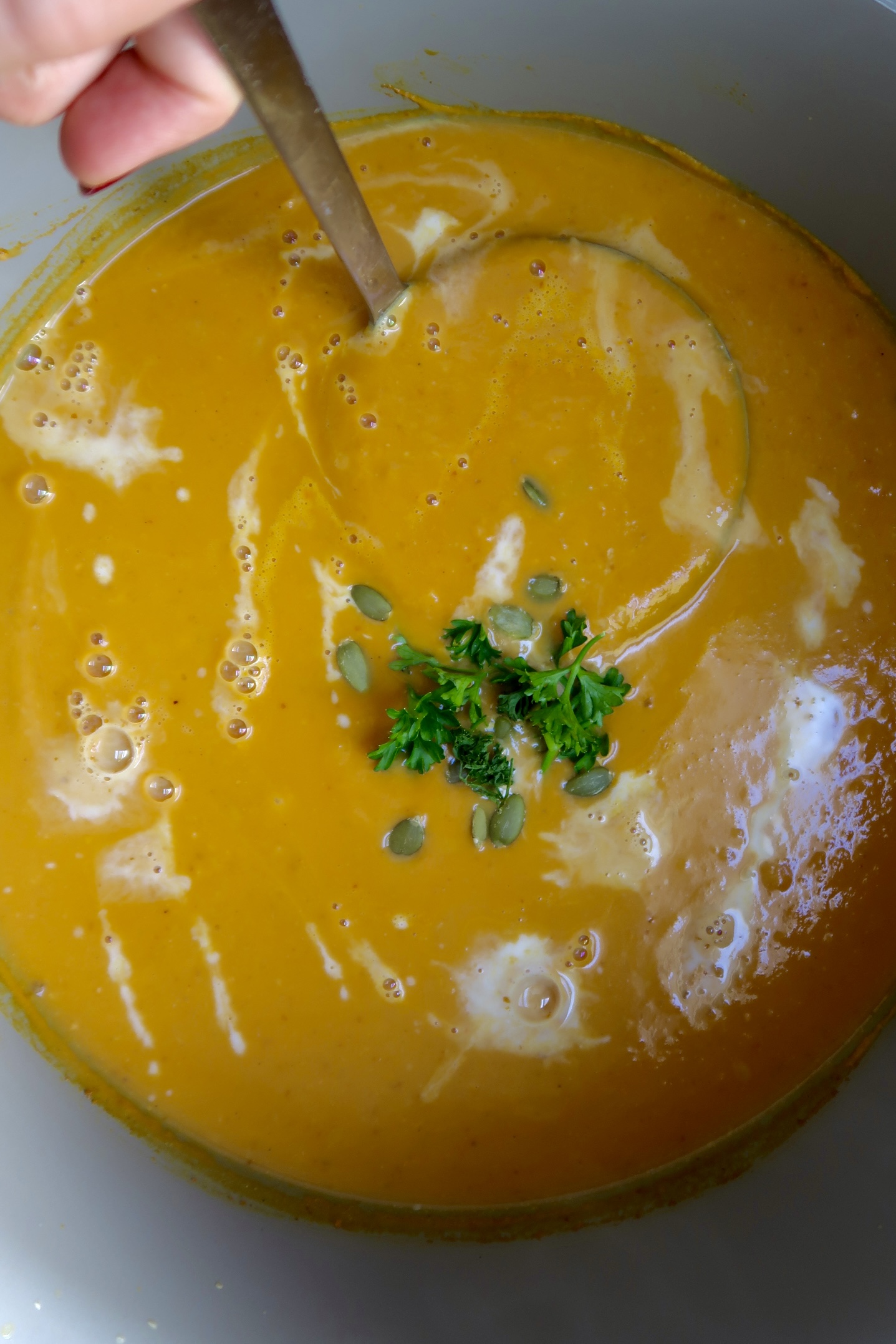Large pot of creamy pumpkin soup with coconut milk drizzled on top throughout and parsley on top and a ladel inside.