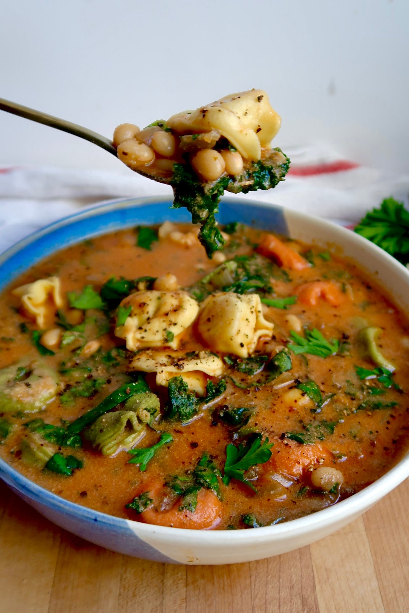 Bowl of soup with tortellini with kale and basil with a spoon scooping some with a tortellini and white beans on it.