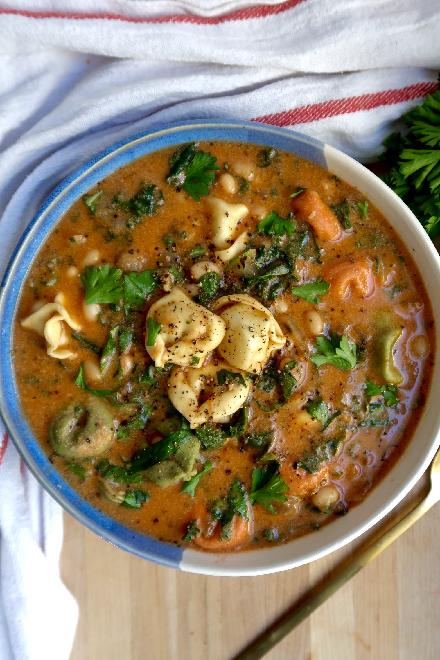 Blue and white bowl with soup with tortellini, kale, basil, and basil off to the side.
