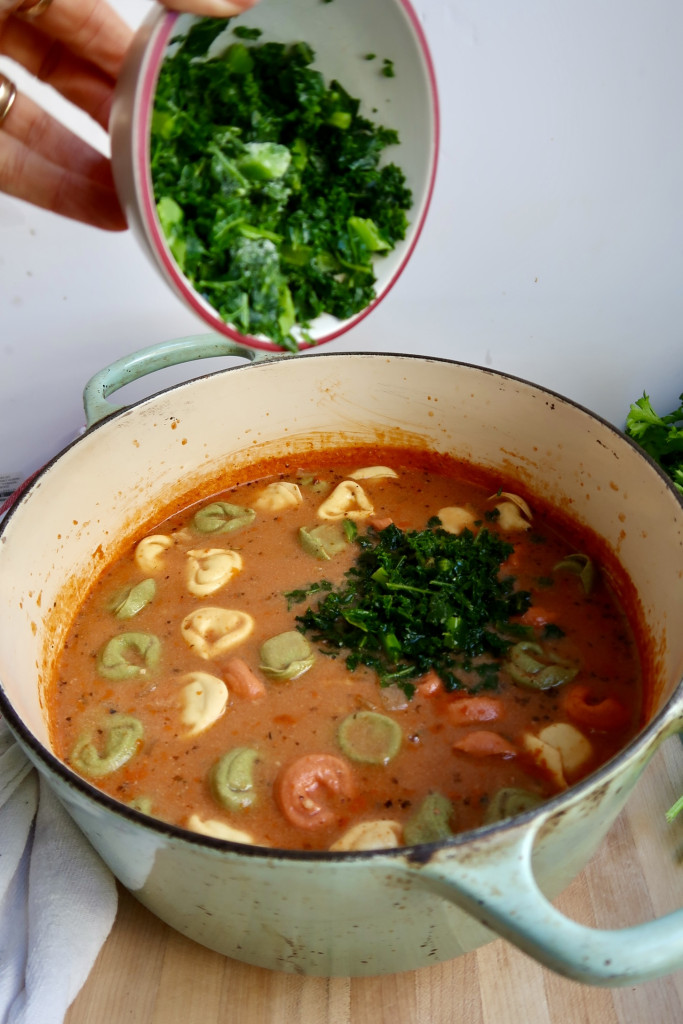 Large pot with tomato soup, tortellini and kale being added to the pot from a small white bowl.