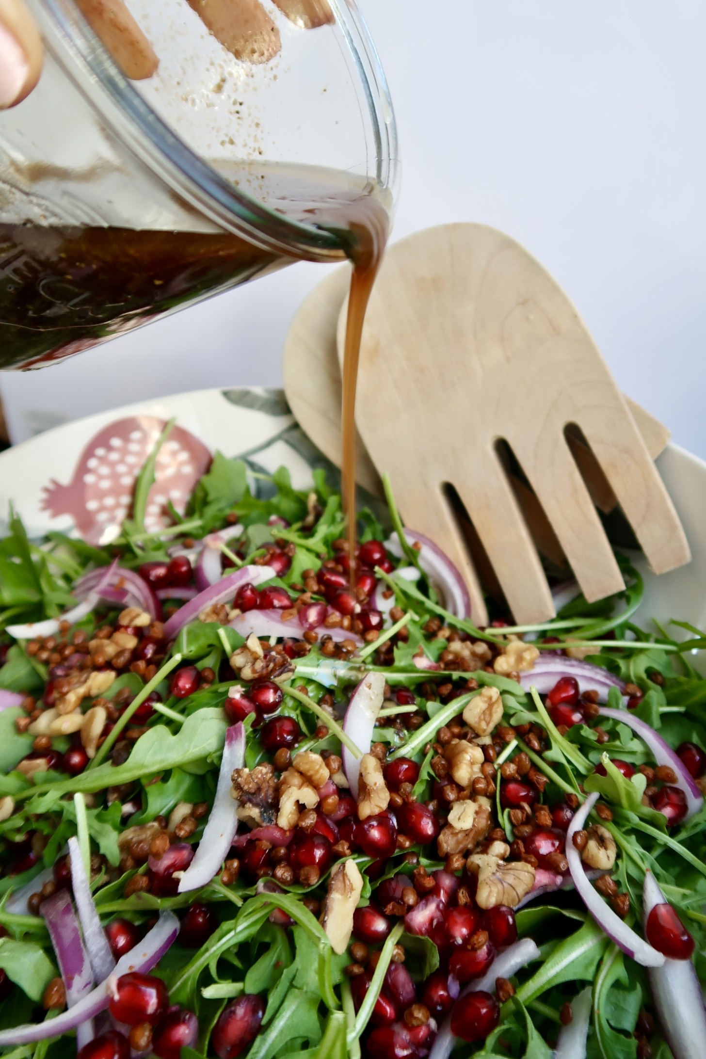 Salad with red onion, pomegranate, walntus and lentils in a bowl with tongs to the side and dressing being poured on top.