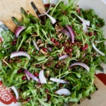 Arugula salad with pomegranate, walnuts, red onion, lentils in it with tongs to the side.