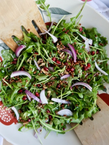 Arugula salad with pomegranate, walnuts, red onion, lentils in it with tongs to the side.