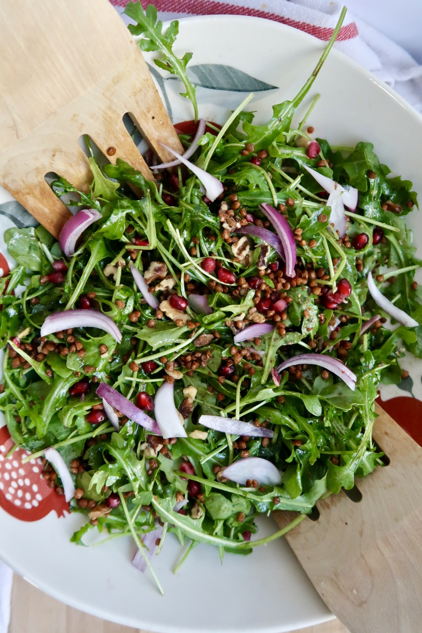 Arugula salad with pomegranate, walnuts, red onion, lentils in it with tongs to the side. 