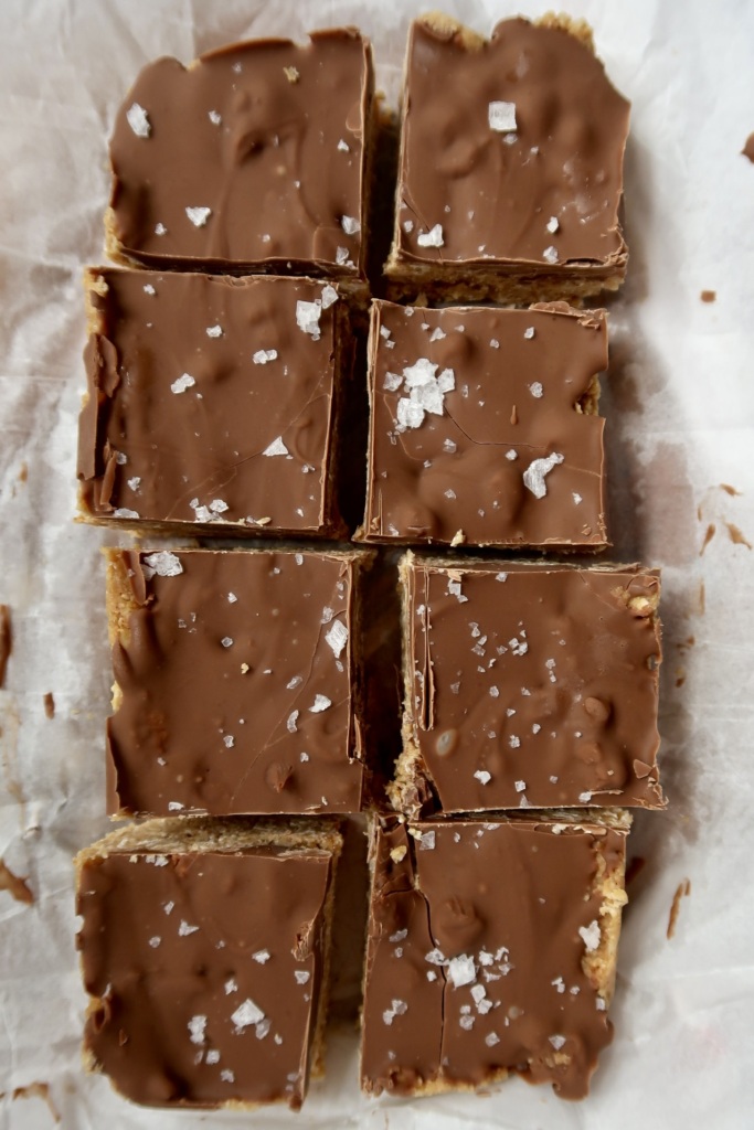 Eight chocolate-covered bars topped with flaky sea salt are arranged in two rows of four on parchment paper. The bars have a slightly crumbly texture visible at the edges.