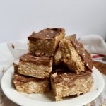A plate stacked with chocolate-topped oat bars. The bars are thick, with a crumbly oat base and a smooth chocolate layer on top, displayed on a white plate against a simple background.