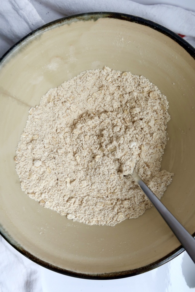 A mixing bowl contains dry ingredients, likely flour and oats, with a metal spoon partially visible on the right side.