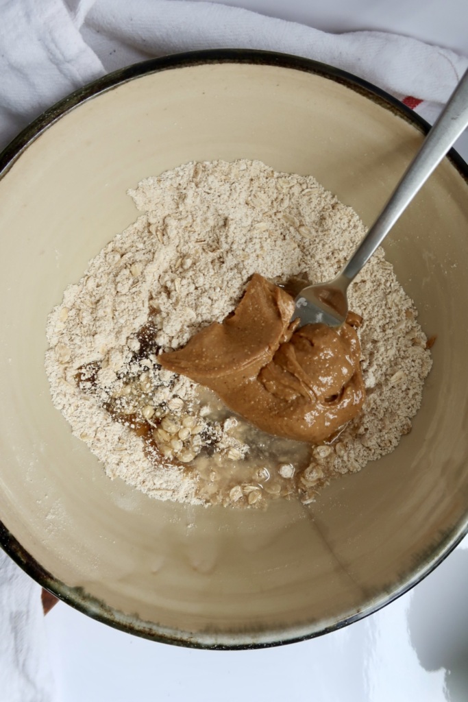 A mixing bowl containing flour, oats, peanut butter, and a fork, with a white towel partially visible underneath.