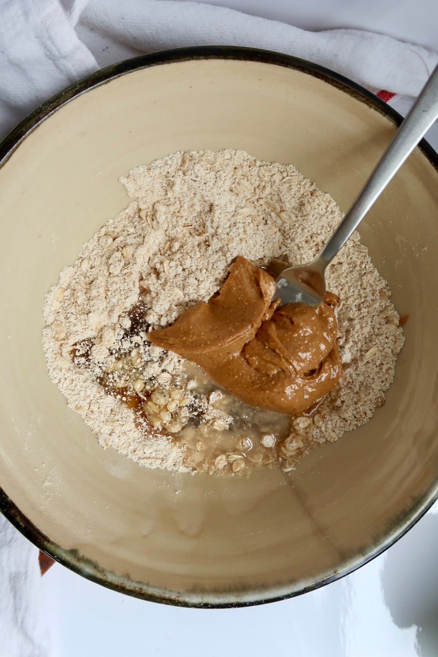 A mixing bowl containing flour, oats, peanut butter, and a fork, with a white towel partially visible underneath.
