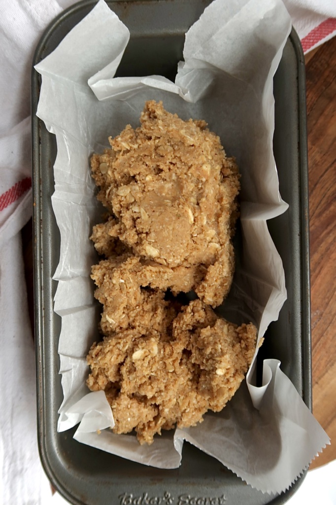 A loaf pan lined with parchment paper holds an unbaked mixture of oats and dough, ready to be pressed and baked. The pan sits on a wooden surface near a white and red cloth.