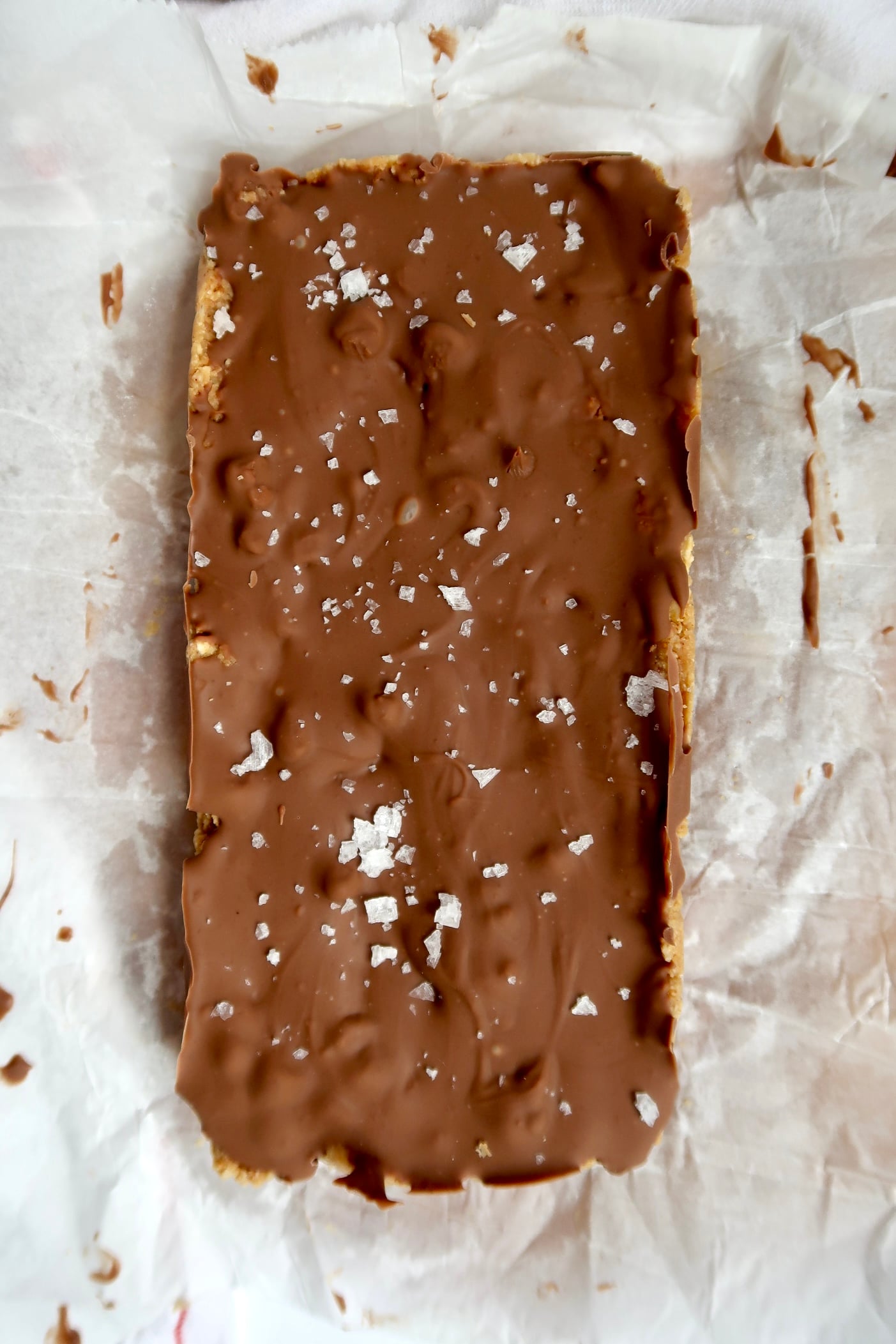 A rectangular chocolate-covered dessert topped with flaky sea salt sits on crumpled parchment paper.