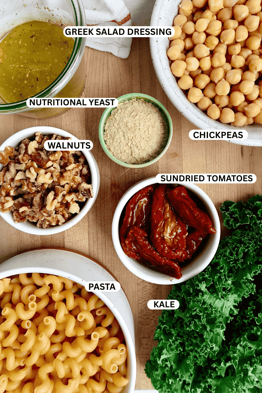 Flatlay of ingredients for pasta salad including kale, chickpeas, sundried tomatoes, pasta, walnuts, nutritional yeast, and salad dressing.
