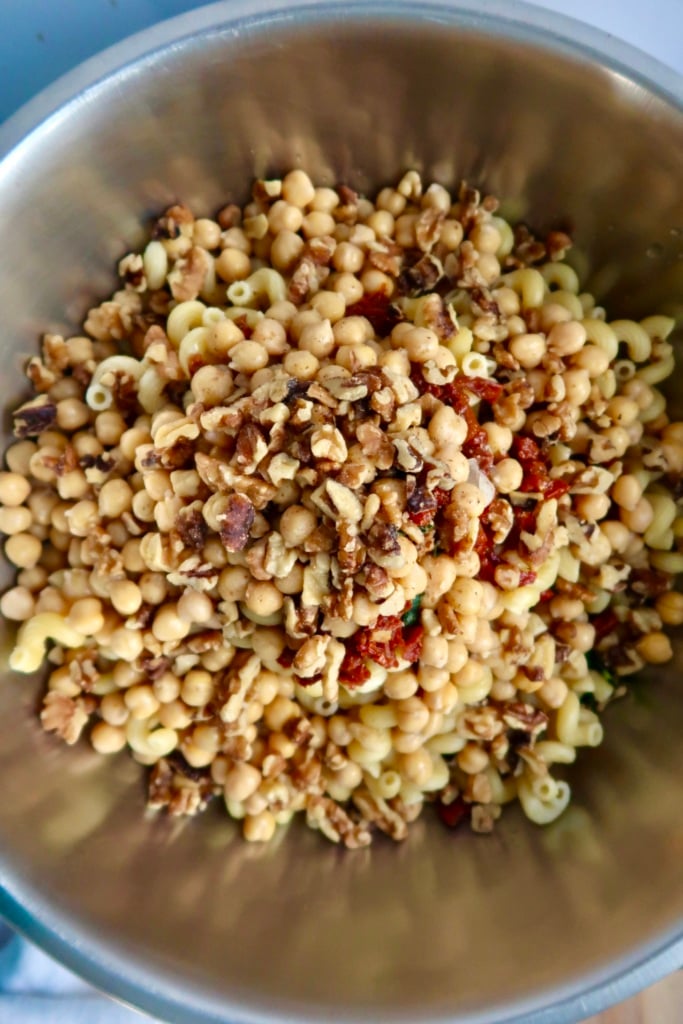 A metal bowl filled with a mixture of elbow macaroni, chickpeas, chopped walnuts, and dried cranberries.