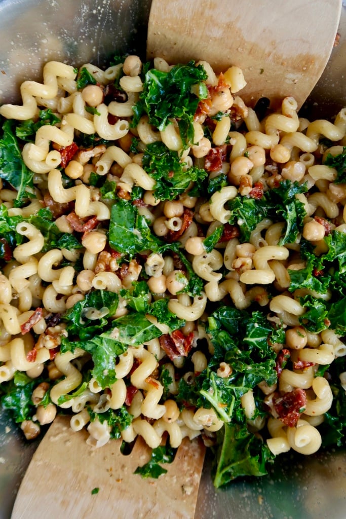 A close-up of pasta salad with curly noodles, kale, chickpeas, sun-dried tomatoes, and herbs being mixed with two wooden utensils in a metal bowl.
