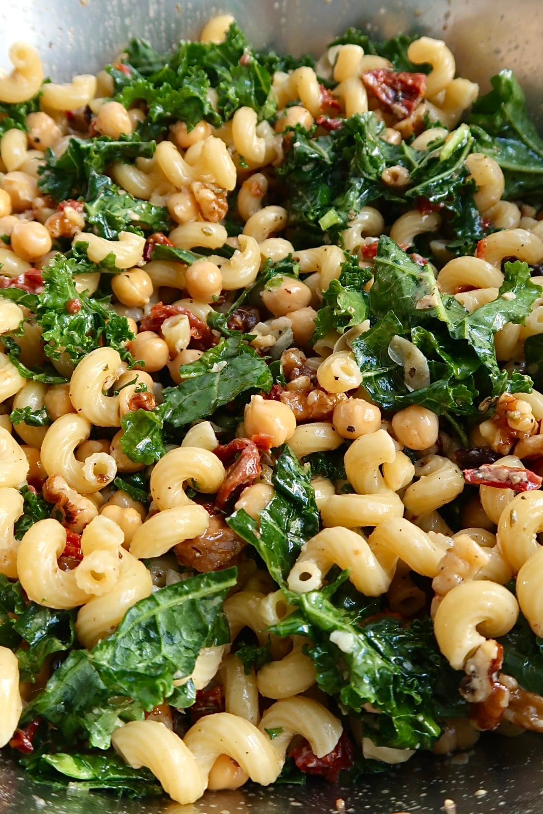 A close-up of pasta salad with curly noodles, chickpeas, sun-dried tomatoes, fresh kale, and a light dressing, all mixed together in a bowl.
