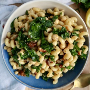 A bowl of curly pasta mixed with kale, chickpeas, and sun-dried tomatoes sits on a blue and white plate, with a gold fork nearby and a white cloth in the background.