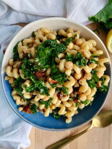 A bowl of curly pasta mixed with kale, chickpeas, and sun-dried tomatoes sits on a blue and white plate, with a gold fork nearby and a white cloth in the background.