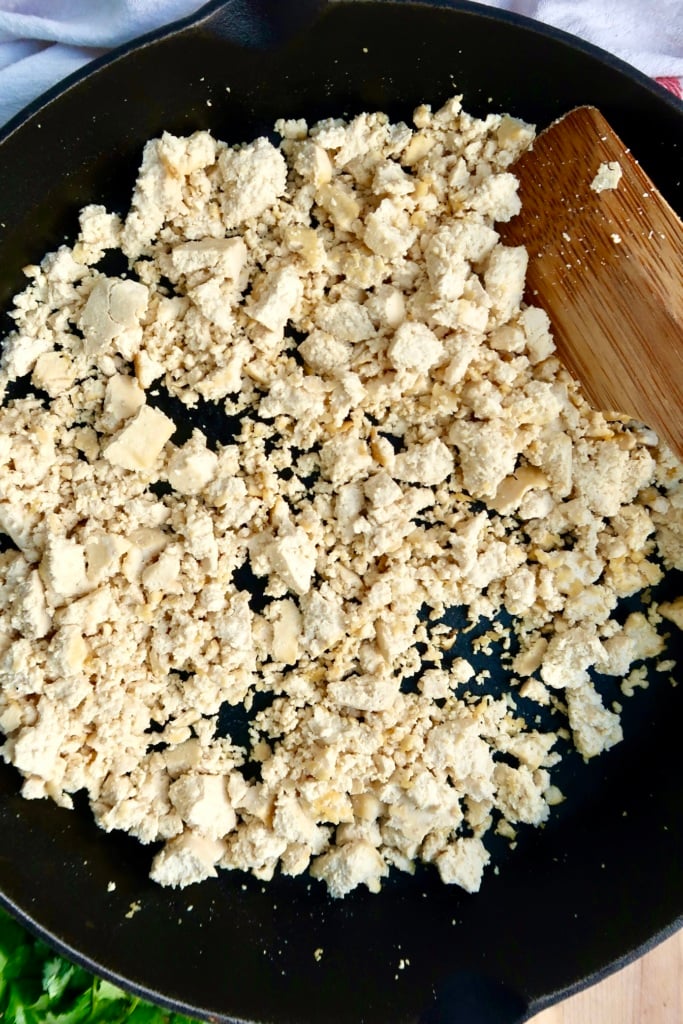 Crumbled tofu cooking in a black skillet, being stirred with a wooden spatula.