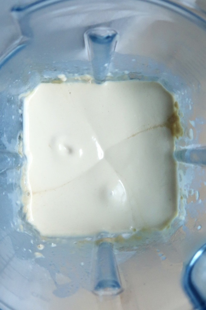 A close-up view of thick, creamy white liquid inside a blender, seen from above. The blender sides are transparent with blueish tint, and some liquid is splattered on the walls.