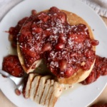 A stack of pancakes topped with strawberry compote and shredded coconut sits on a white plate. A fork with a sliced piece of pancake rests beside the stack, with extra compote on the plate and table.