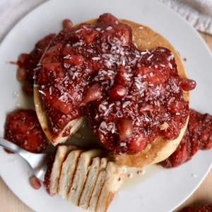 A stack of pancakes topped with strawberry compote and shredded coconut sits on a white plate. A fork with a sliced piece of pancake rests beside the stack, with extra compote on the plate and table.