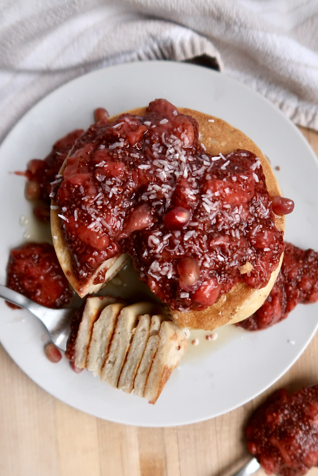 A stack of pancakes topped with strawberry compote and shredded coconut sits on a white plate. A fork with a sliced piece of pancake rests beside the stack, with extra compote on the plate and table.