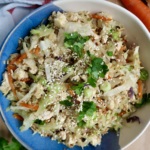 A bowl of stir-fried shredded cabbage, carrots, green onions, and tofu, topped with sesame seeds and fresh cilantro, sits on a wooden surface with a sprig of cilantro, ginger, and a carrot nearby.