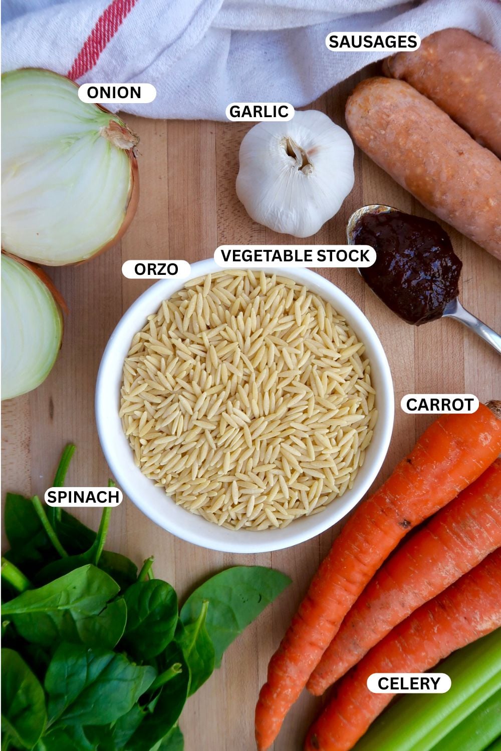 A bowl of orzo pasta surrounded by labeled ingredients: onion, garlic, sausages, vegetable stock, carrot, celery, and spinach, all arranged on a wooden surface.