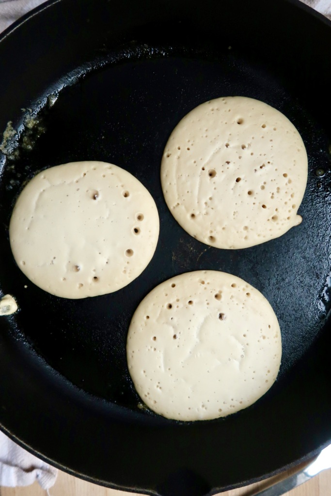 Three pancakes cooking in a black skillet, with bubbles forming on the surface, indicating they are almost ready to be flipped.