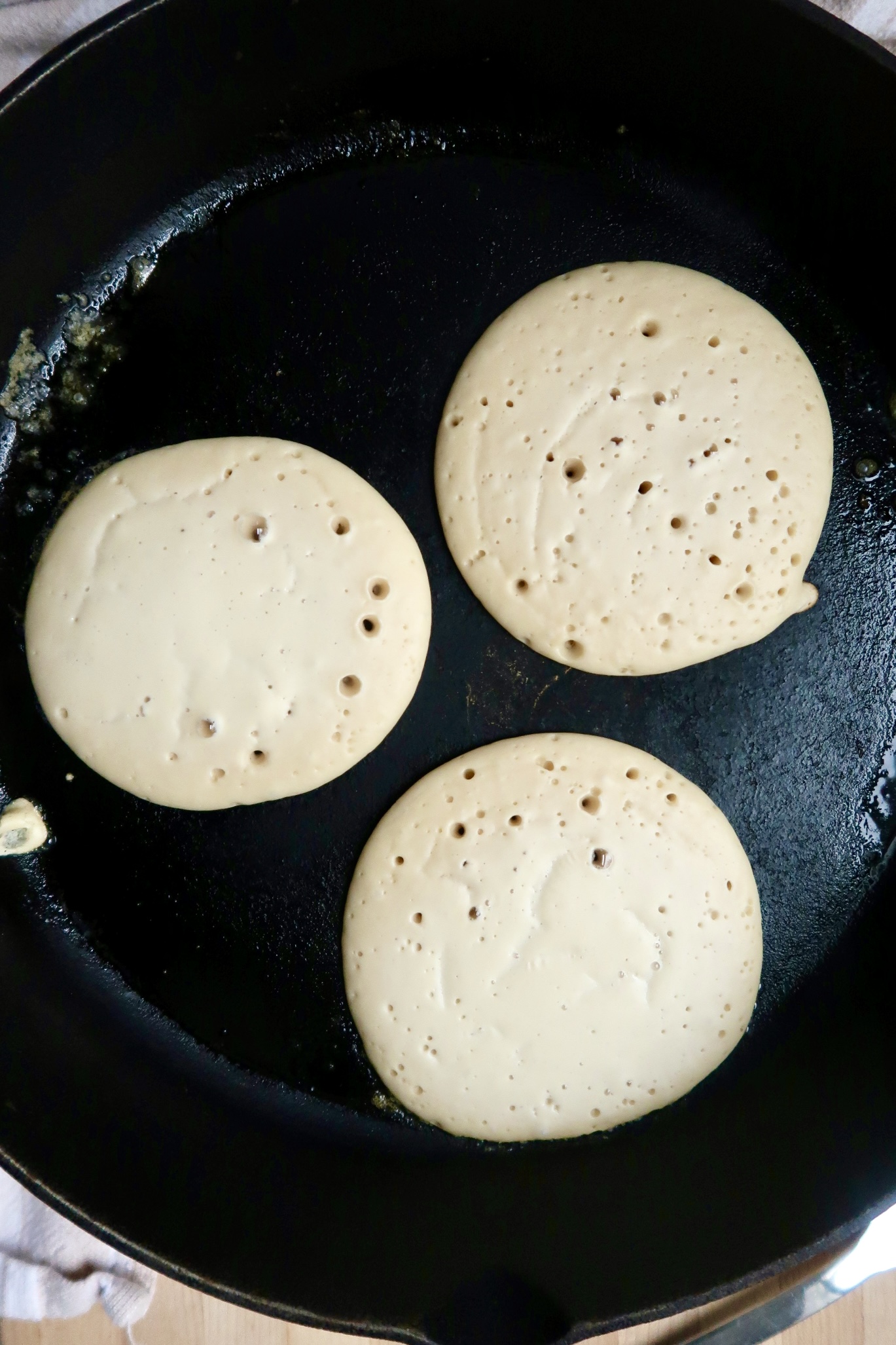 Three pancakes cooking in a black skillet, with bubbles forming on the surface, indicating they are almost ready to be flipped.