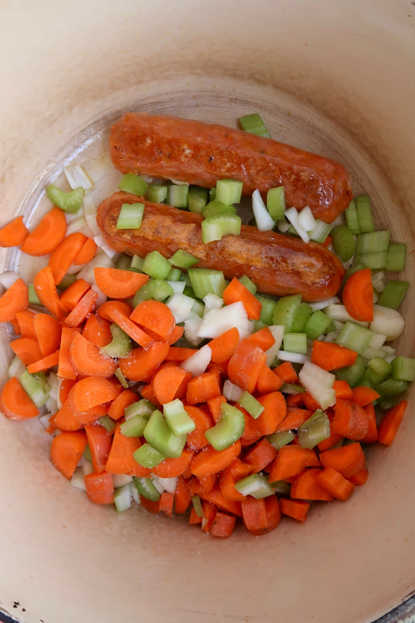 Two sausages in a pot with chopped carrots, celery, and onions, ready to be cooked.